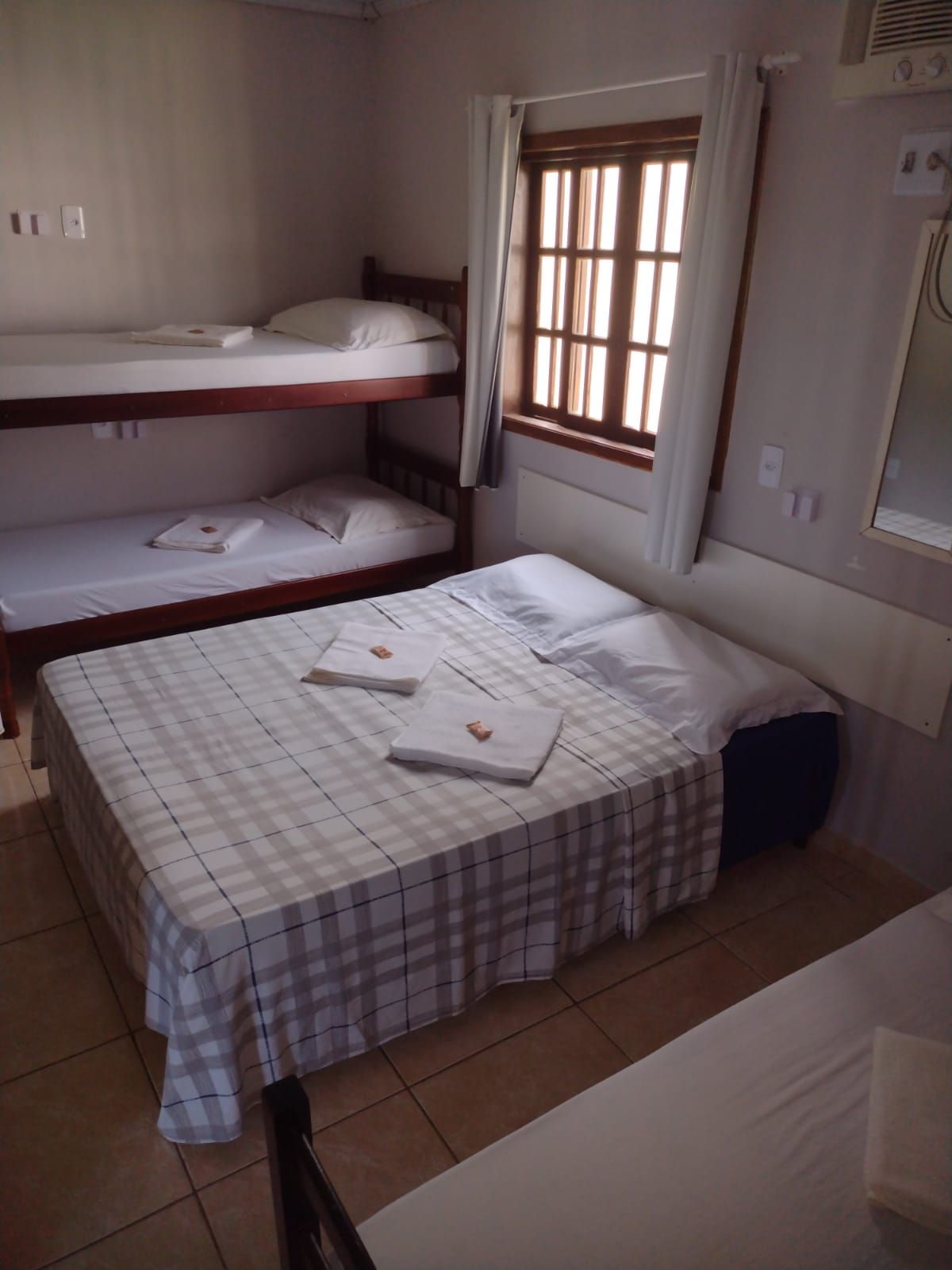 Um quarto de hotel com uma cama de casal coberta com uma manta xadrez, um beliche de madeira e uma janela com cortinas brancas.