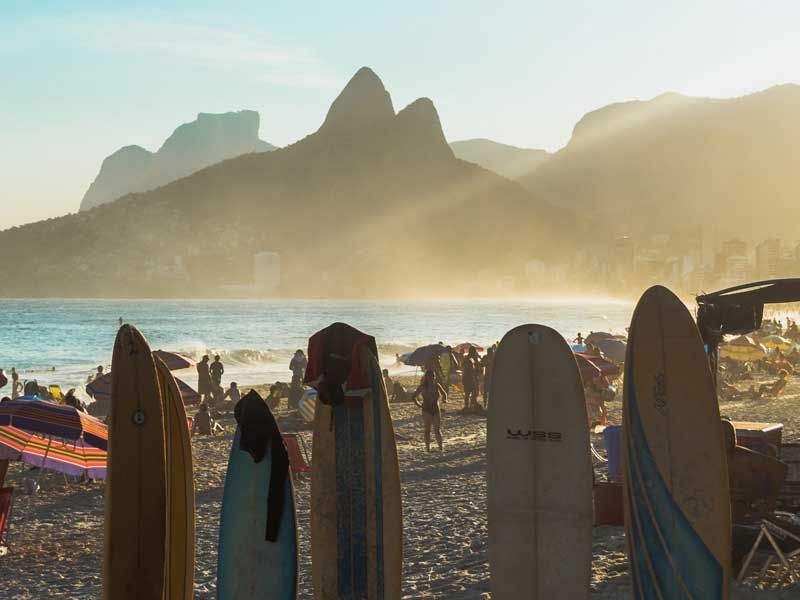Pranchas de surfe permanecem em pé na areia da praia ao pôr do sol, com a icônica Serra Dois Irmãos ao fundo.