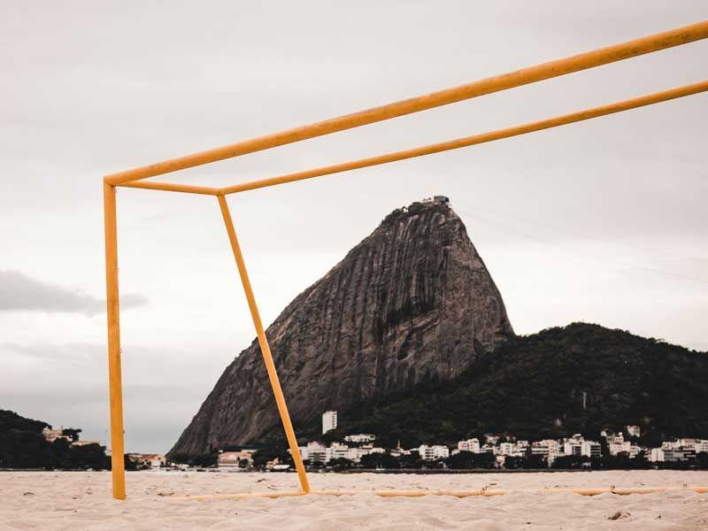 Um gol de futebol amarelo em uma praia de areia, com o Pão de Açúcar ao fundo sob um céu nublado.