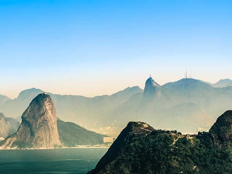 O Pão de Açúcar se ergue acima de uma baía no Rio de Janeiro, com a estátua do Cristo Redentor visível em um pico distante.