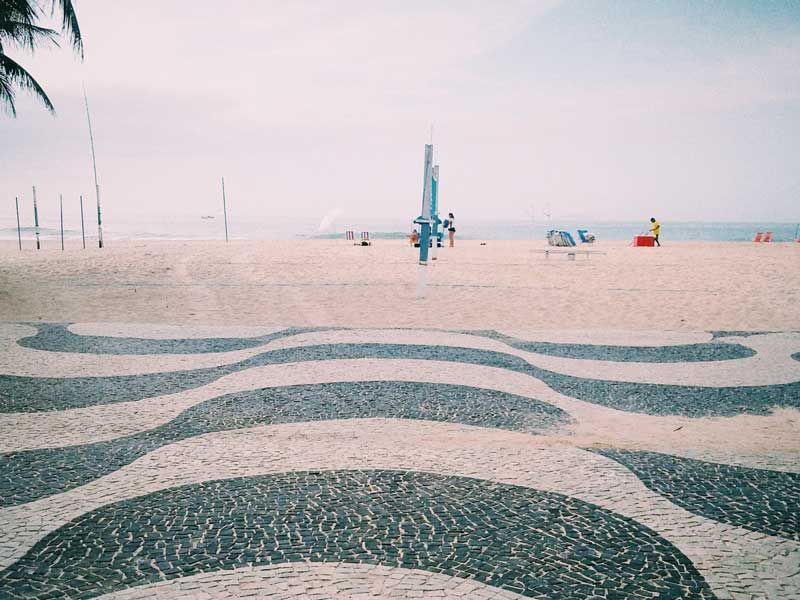 Calçada icônica com padrão de ondas em preto e branco na praia de Copacabana, que leva à areia e ao oceano.
