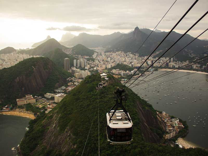 Um teleférico percorre uma colina verdejante em direção à cidade litorânea do Rio de Janeiro, com montanhas ao fundo.