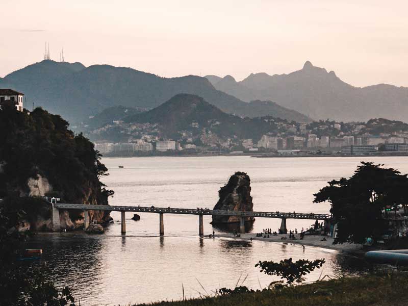 Uma longa ponte pedonal liga uma costa rochosa a uma pequena ilha ao pôr do sol, com montanhas ao fundo.