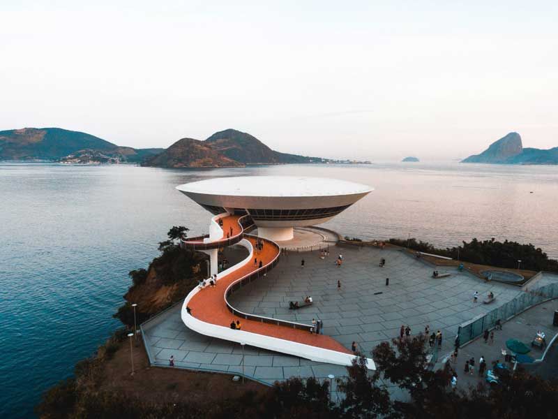 O Museu de Arte Contemporânea de Niterói, um edifício branco em forma de pires, ergue-se numa falésia com vista para o mar, acessível por uma rampa curva.