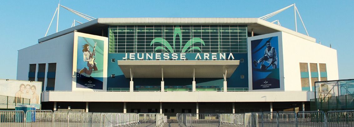 A Jeunesse Arena é um grande estádio moderno com fachada branca, centro envidraçado e dois cartazes de destaque em cada lado.
