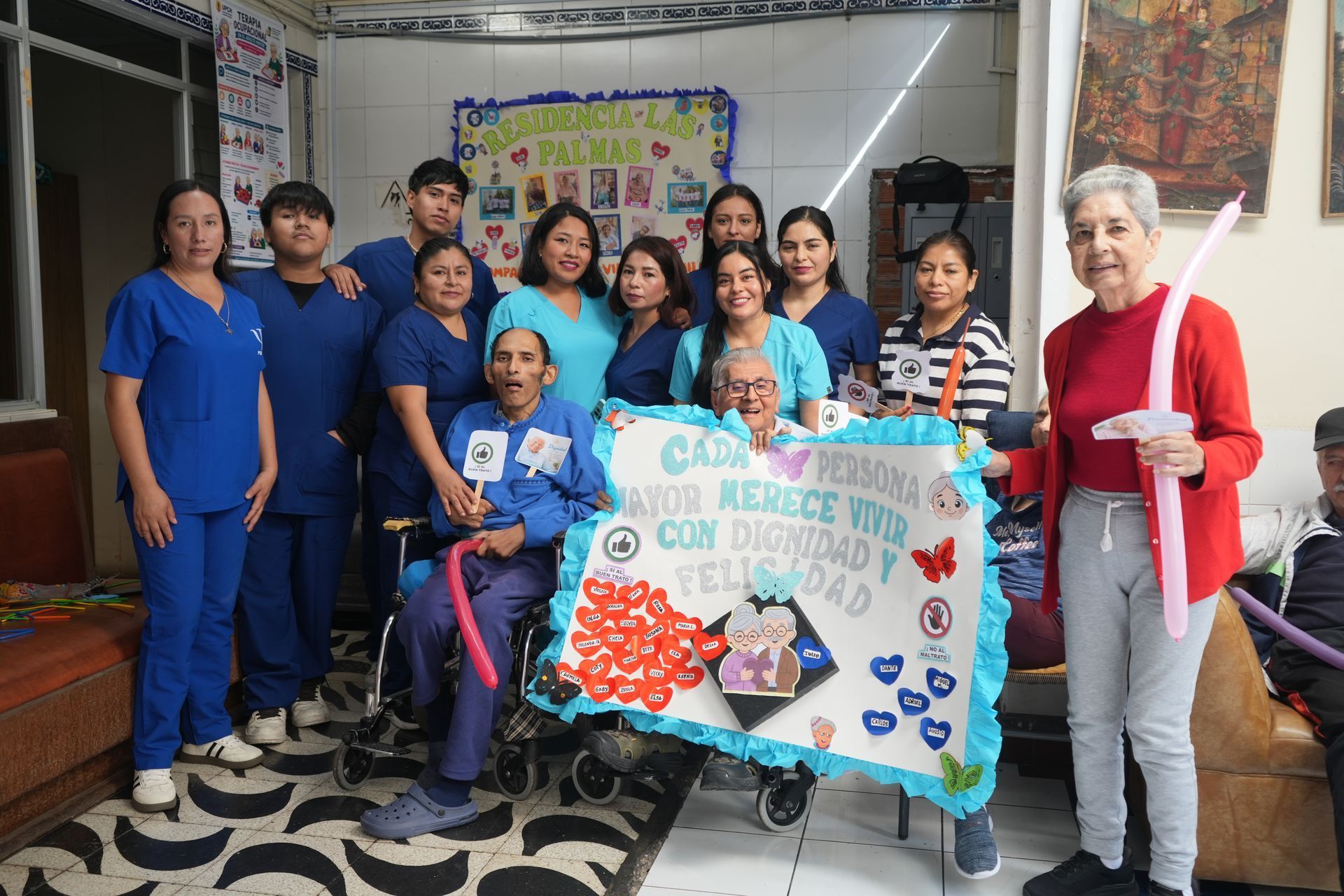 Grupo sosteniendo un cartel hecho a mano y globos durante una celebración comunitaria en el interior.
