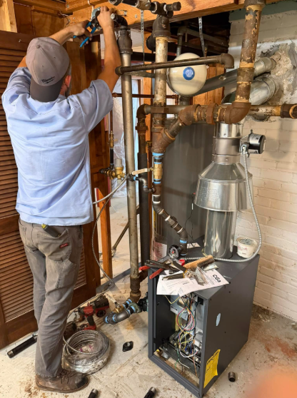 Man in light blue shirt working on plumbing in a utility room. Boiler and pipes are visible.