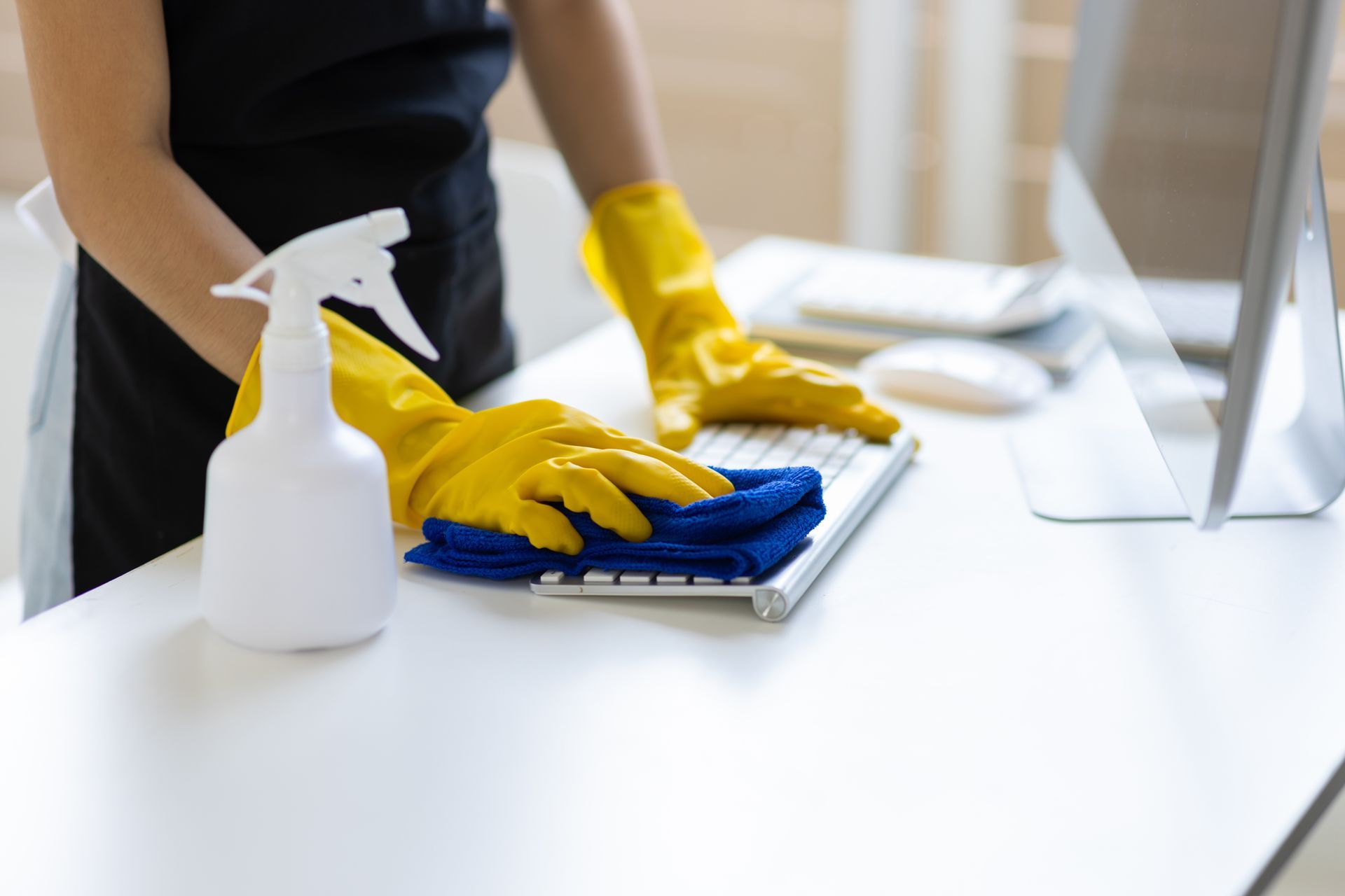 Person in yellow gloves cleans a keyboard with a blue cloth and spray bottle on a white desk.