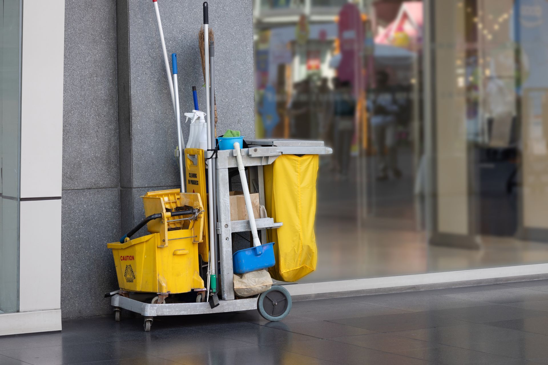 A cleaning cart with a yellow bucket , mop , and broom is parked in front of a building.