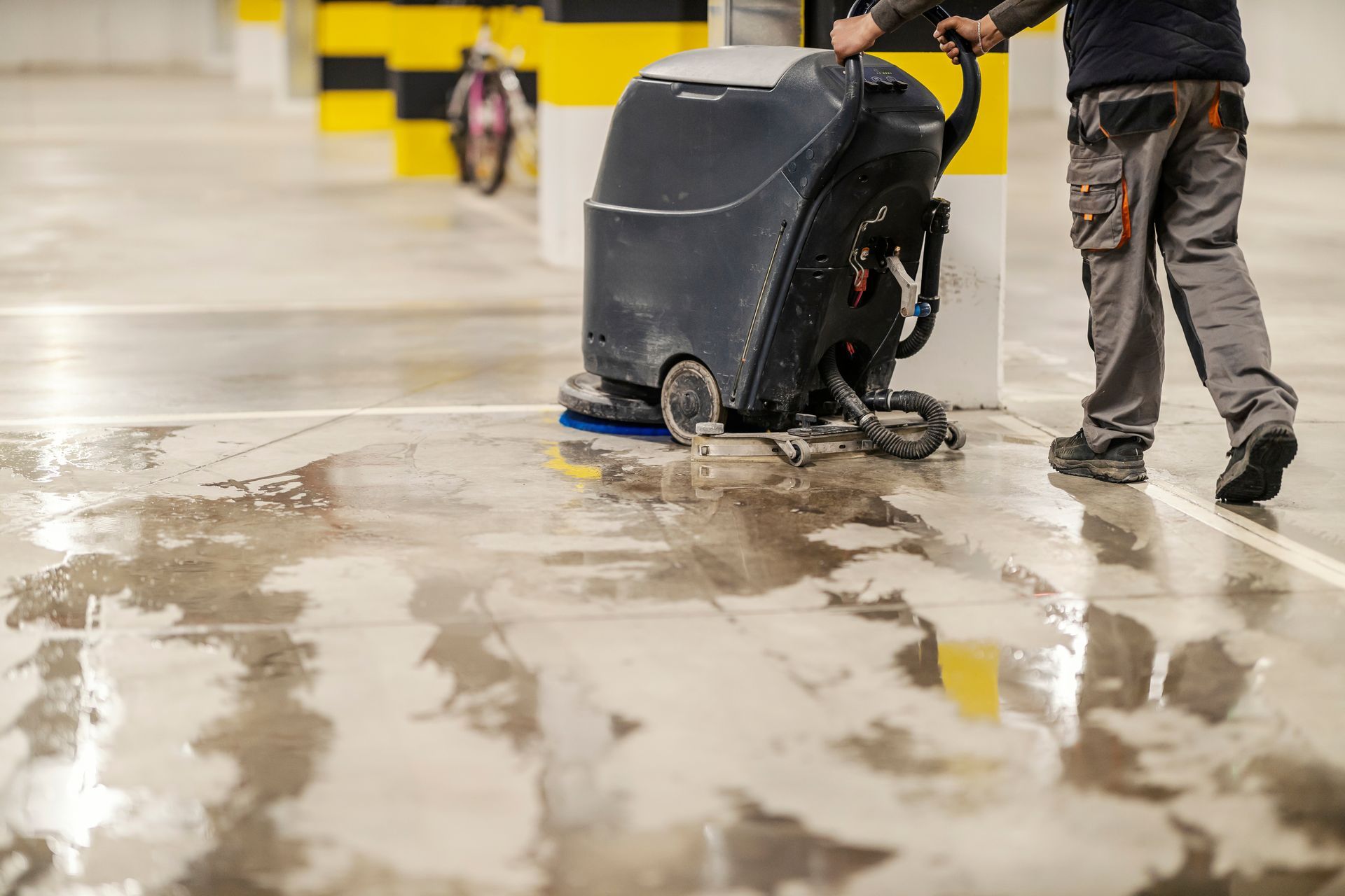 Person operating a floor cleaning machine in a parking garage. Wet floor, concrete, yellow and black pillars.