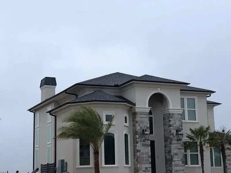 Two-story stucco house with a dark roof and stone accents, palm trees, and an overcast sky.