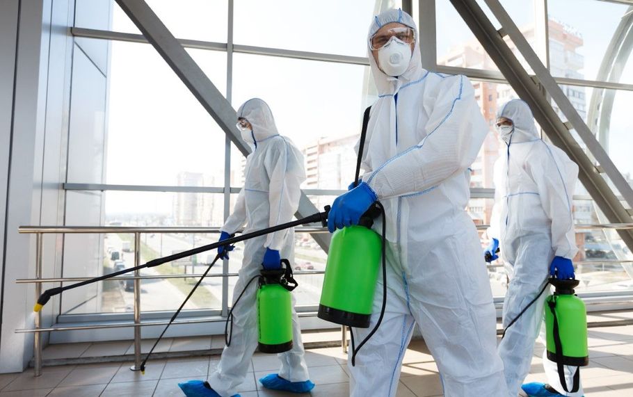 Three workers in white protective suits spray disinfectant in a bright indoor space.