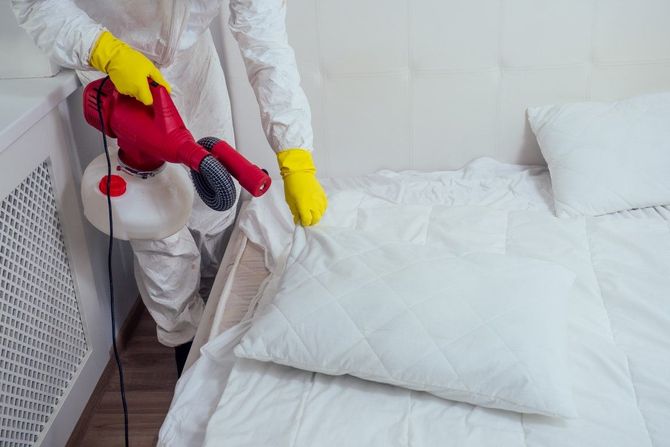Worker in white protective gear spraying a bed with a red disinfectant fogger in a white room