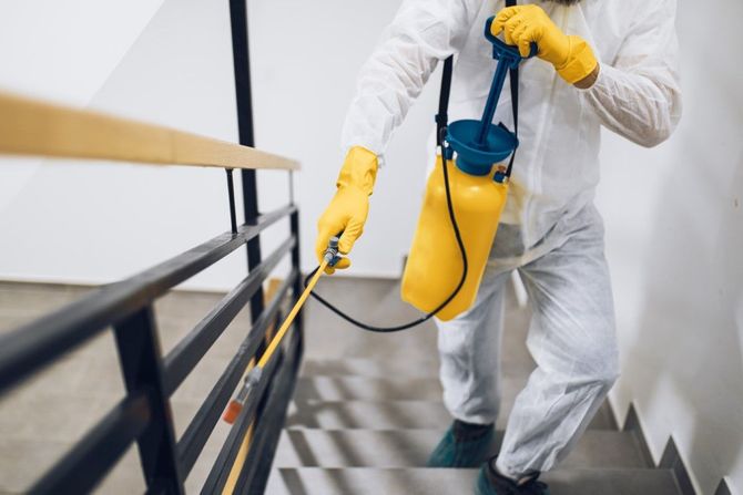 Worker in white protective suit spraying disinfectant on a stairway with a yellow backpack sprayer.