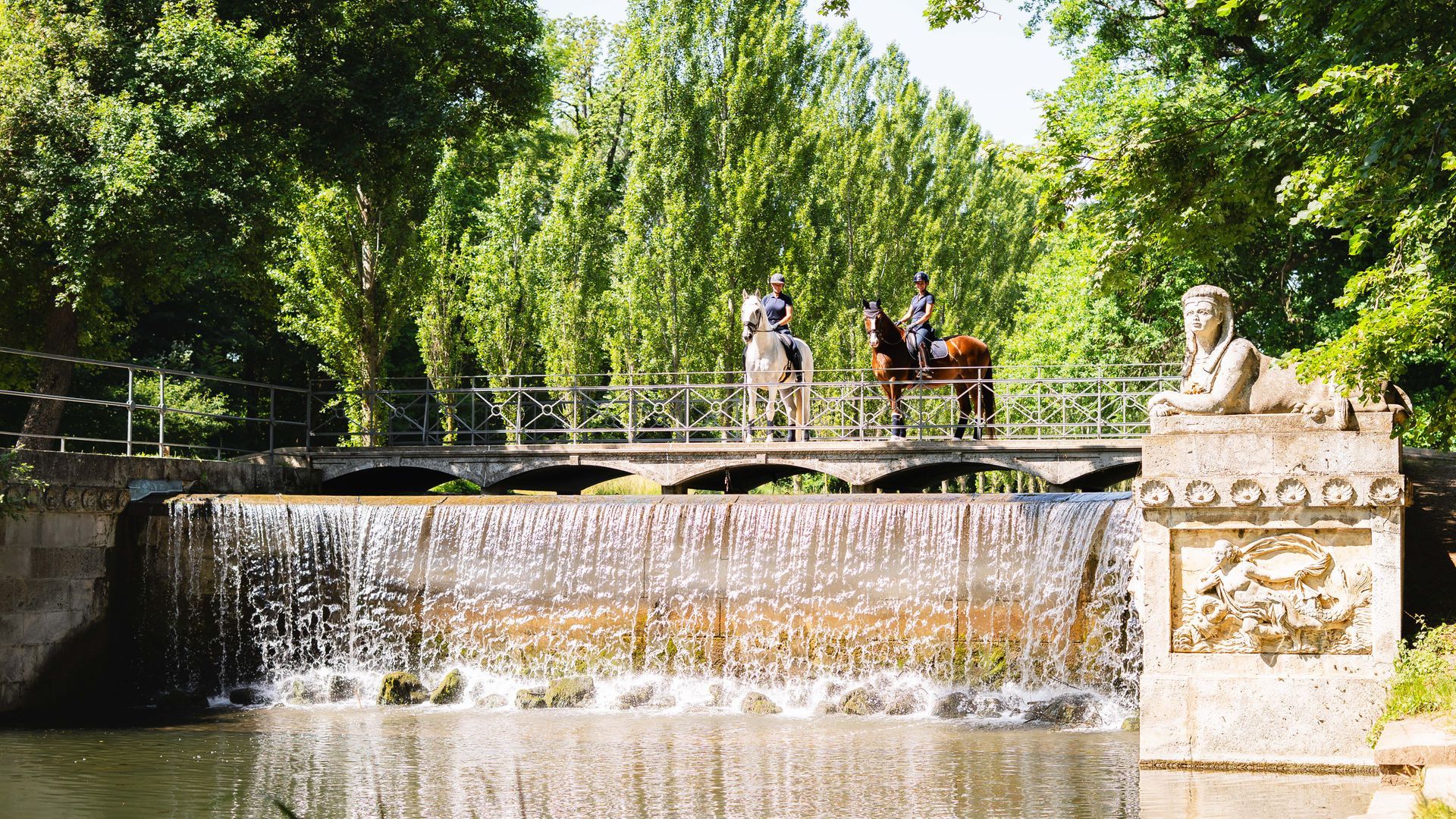 Menschen auf einer Brücke über einem kleinen Wasserfall in einem sonnigen grünen Park