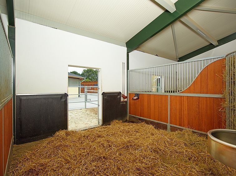 Überdachter Tierstall mit Stroheinstreu, Metallverkleidung und offener Tür zum Hof.