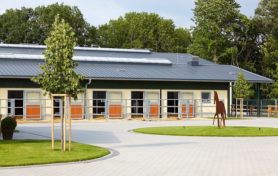 Modernes Stallgebäude mit Solarpaneelen, orangefarbenen Türen und einem gepflasterten Innenhof mit Baum und einer Skulptur