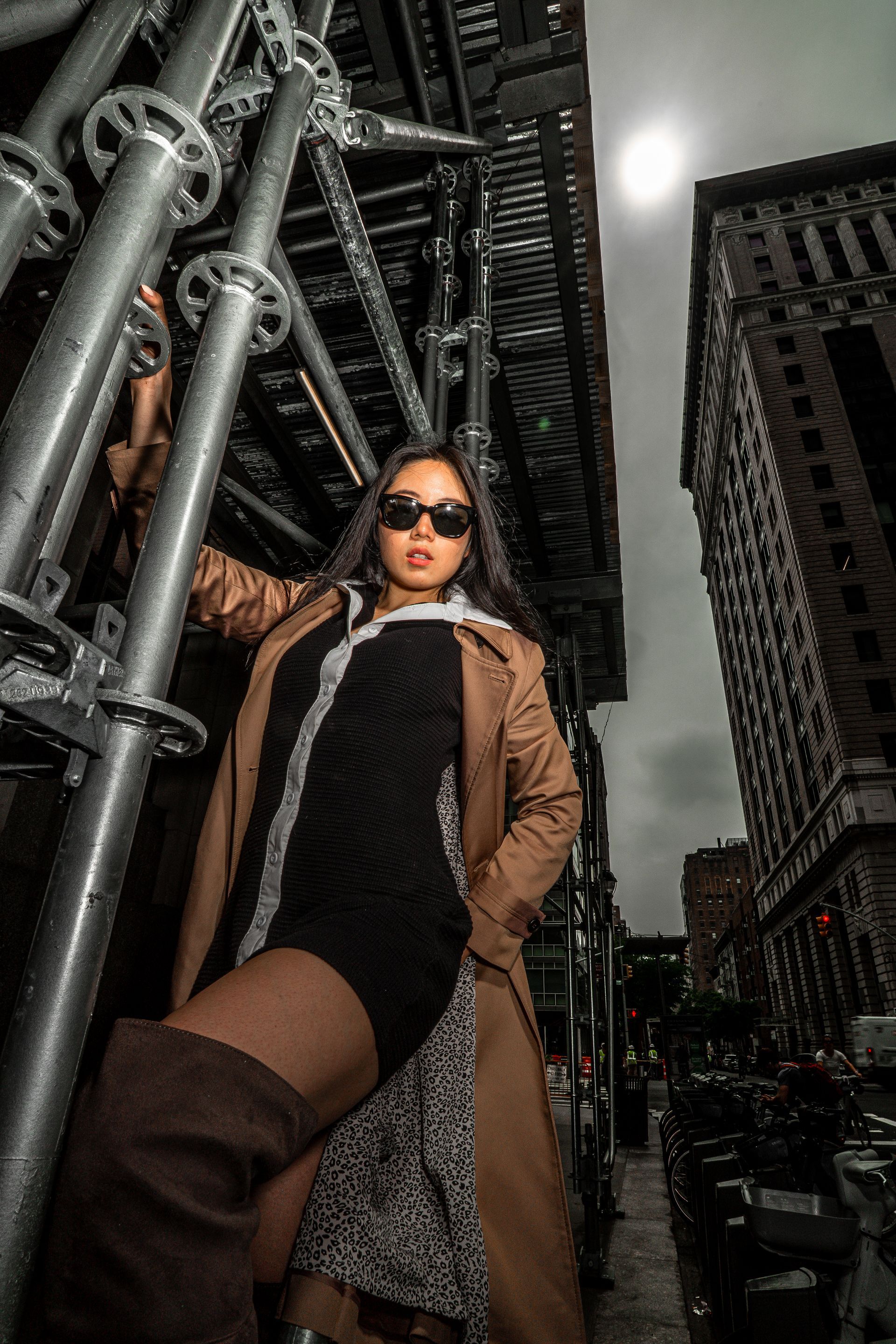 Woman in sunglasses and coat poses by scaffolding on a NYC street under scaffolding. 