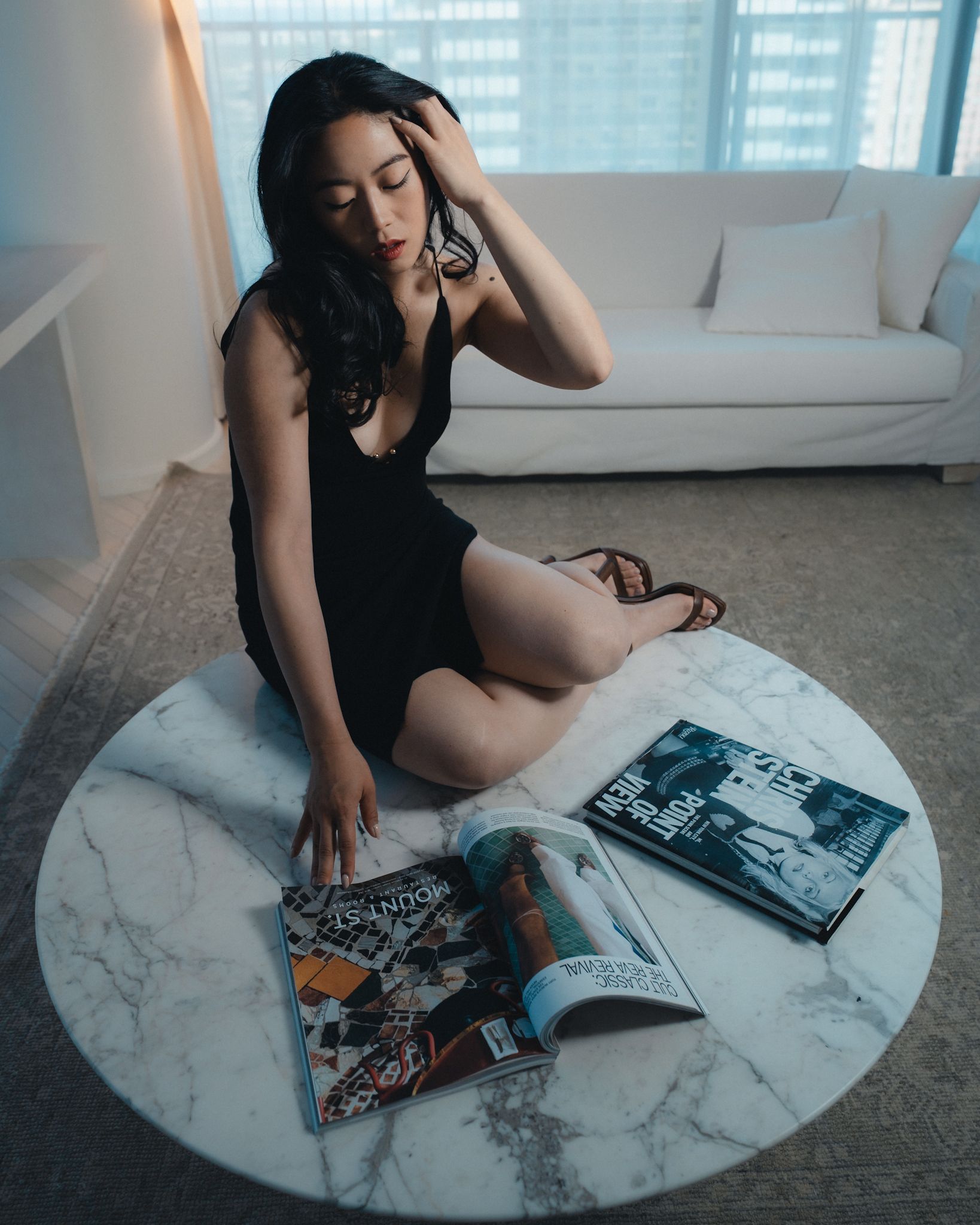 Woman in black dress sits on a marble coffee table with magazines, hotel room setting.