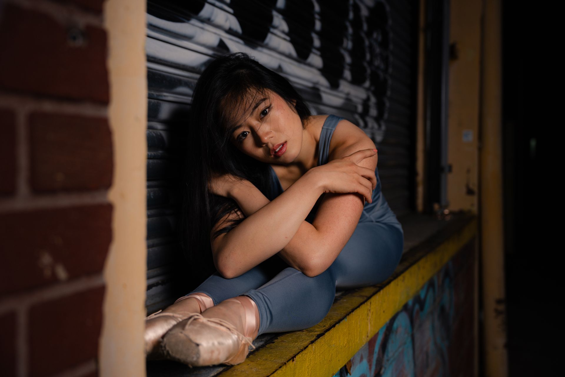 Young Asian woman in ballet attire, sitting on a ledge in an alley, hugging herself, sad expression.