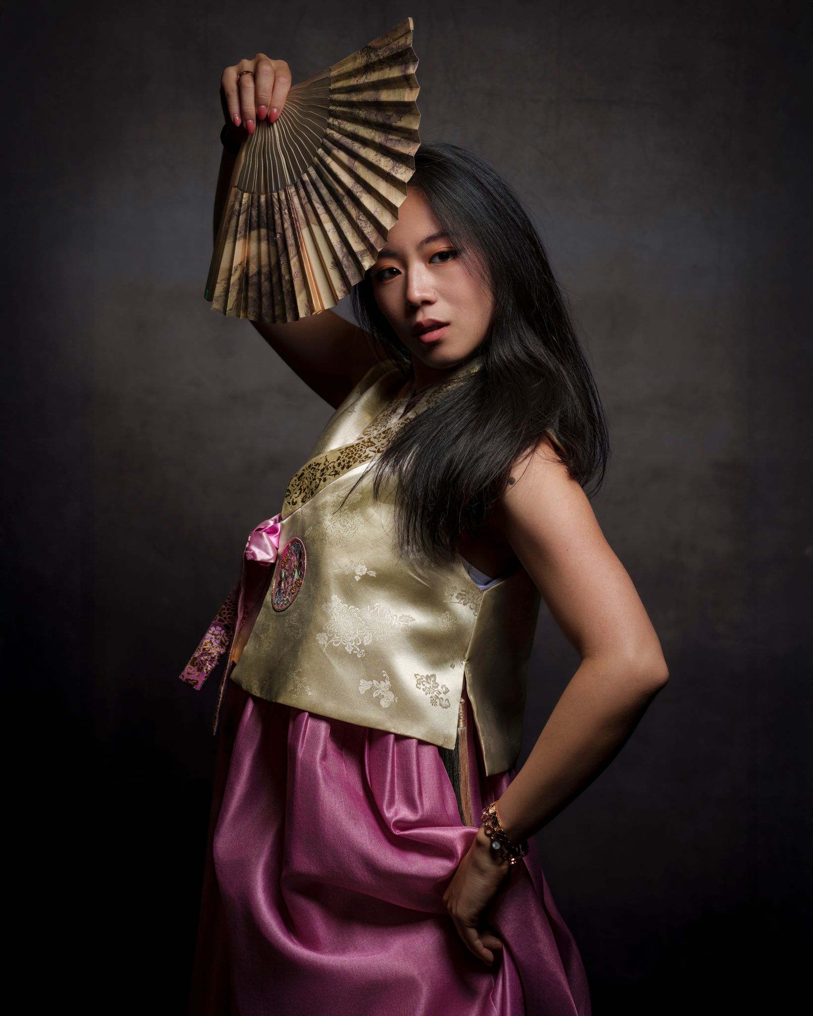 Woman in traditional Korean dress hanbok holding a fan, looking at the viewer, against a dark background.