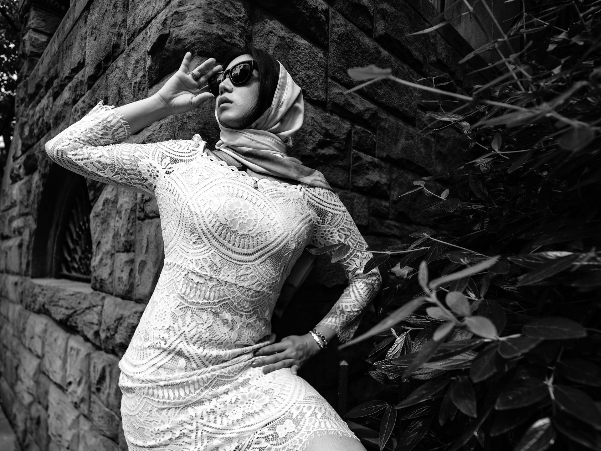 Woman in white lace dress and headscarf poses outdoors, shielding eyes, with stone wall and foliage. Vintage fashion.