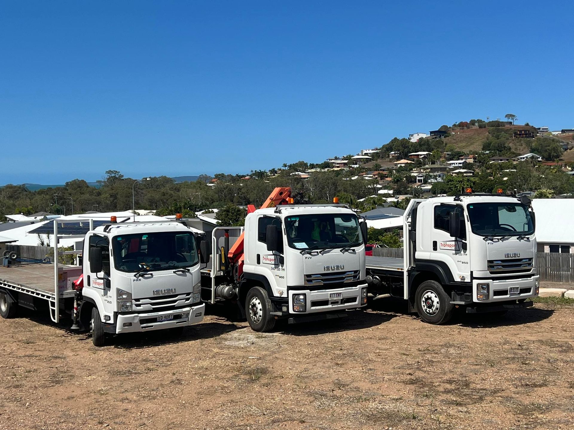 Three white flatbed trucks parked outside under a blue sky, hills in the background.— J & M Transport Plus in Barmaryee, QLD