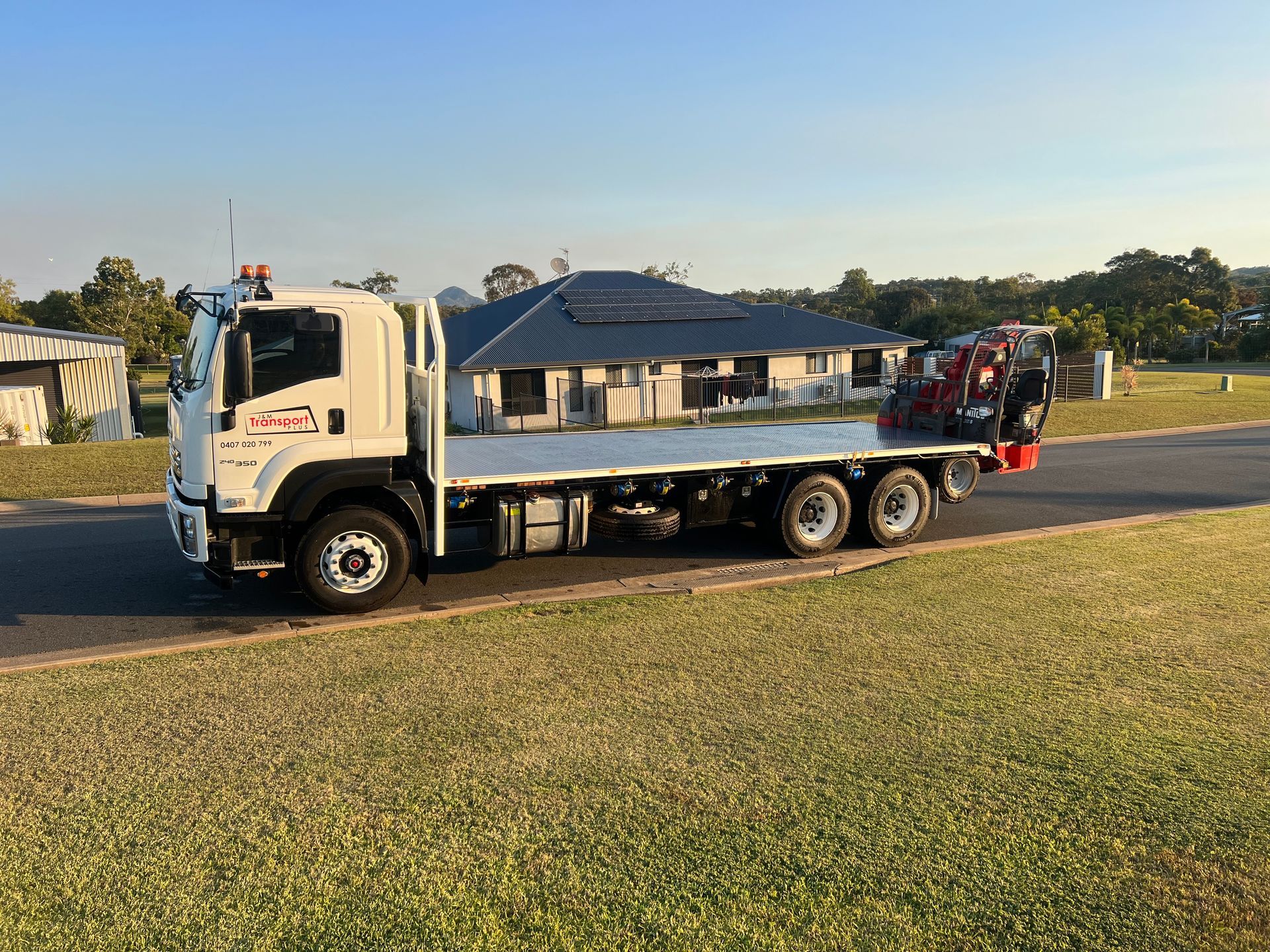 White flatbed tow truck parked on grass beside a road, carrying a red vehicle, with houses in the background