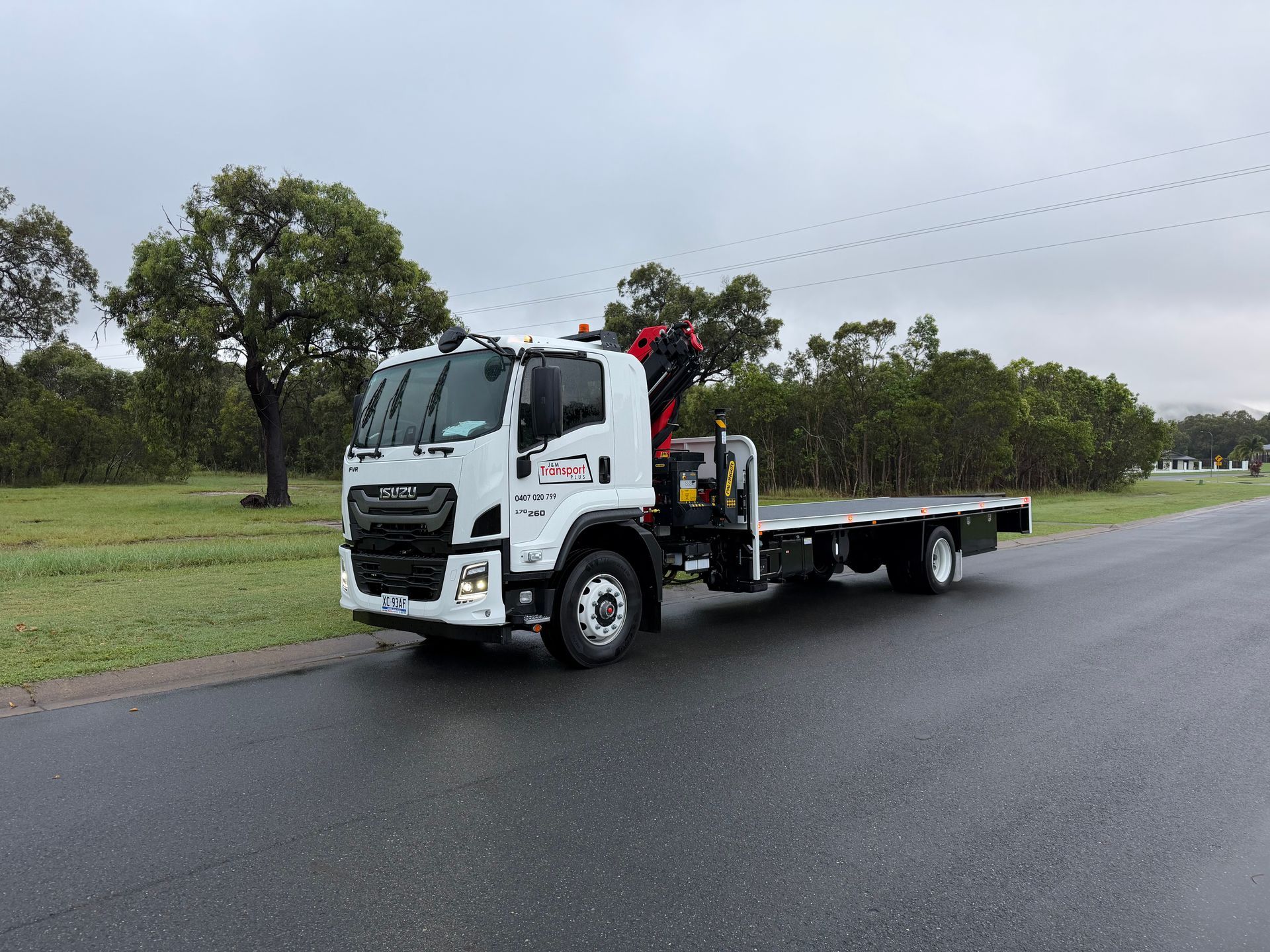 White flatbed tow truck parked on a wet road beside a grassy field and trees under cloudy skies