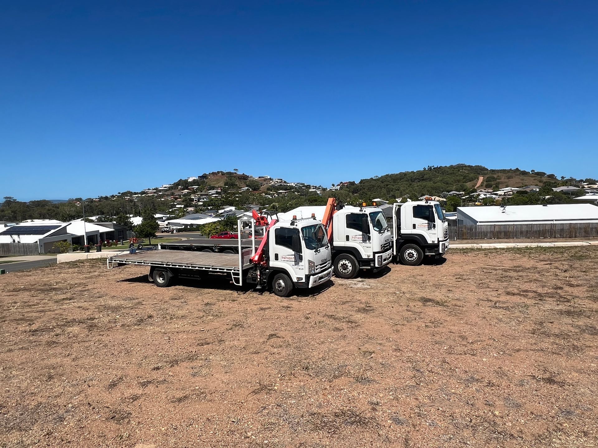 Three white flatbed trucks parked on a dirt lot with hills under a clear blue sky.
