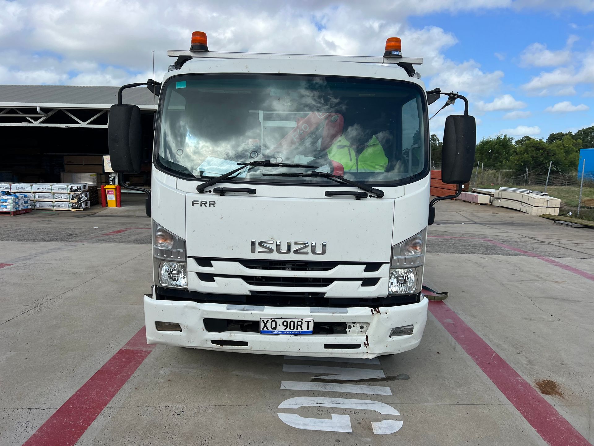 White Isuzu truck parked at a loading bay with orange warning lights and a visible front license plate.