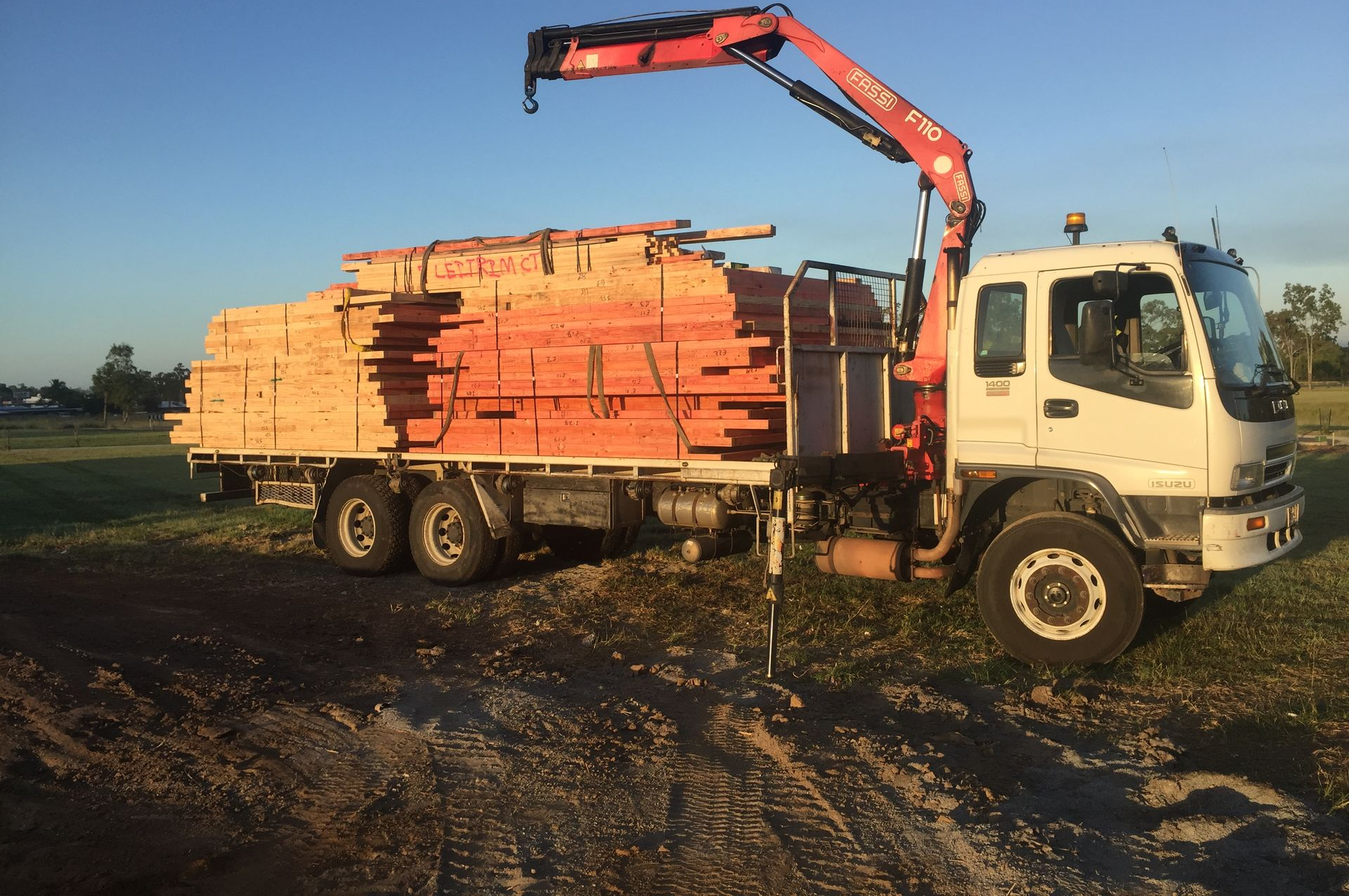 Truck loaded with stacked lumber, with a red crane lifting boards in a muddy field at sunset