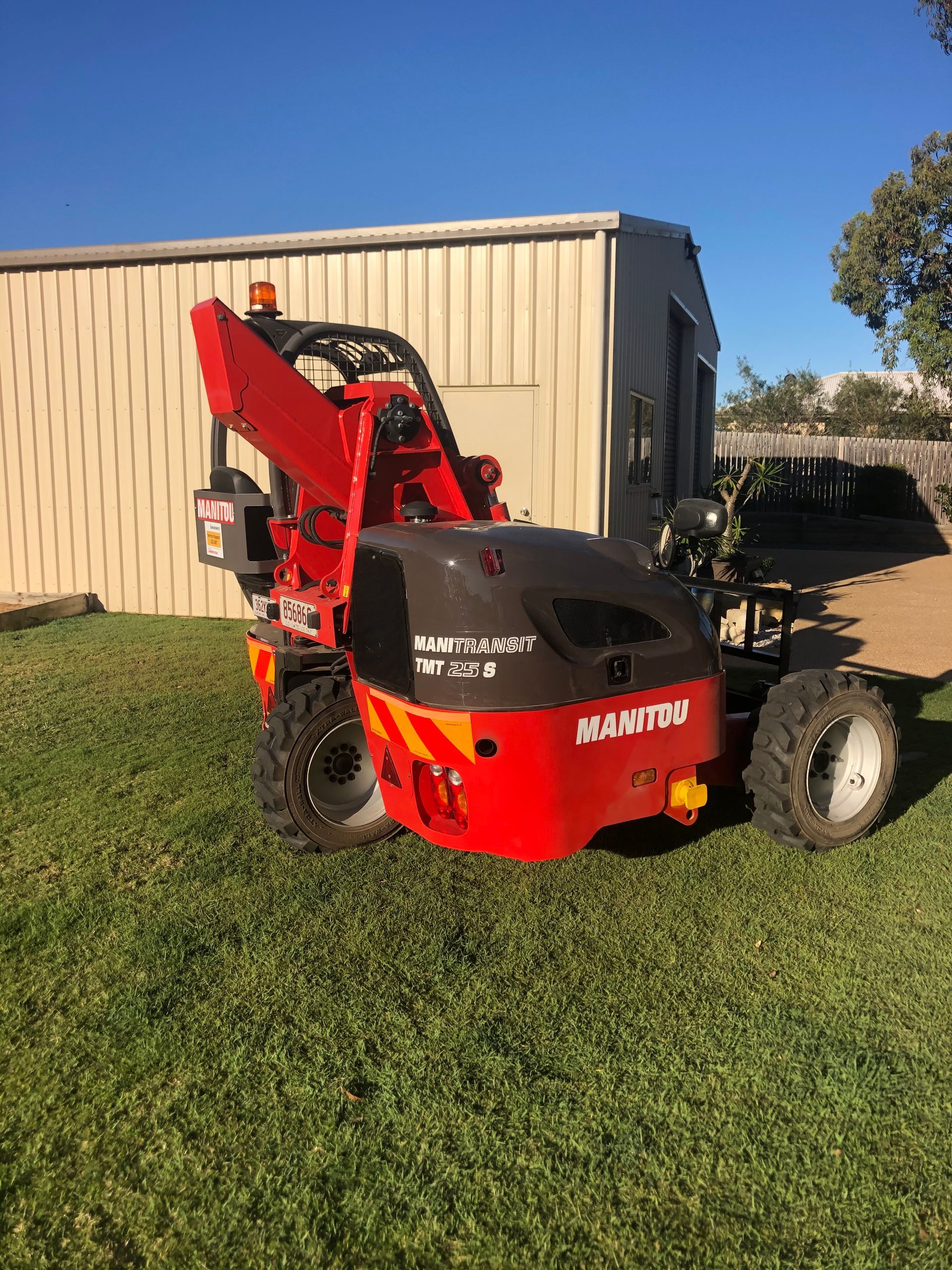 Red compact loader parked on grass beside a beige metal shed in sunlight