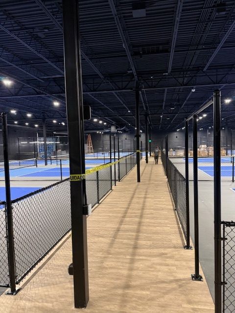 Indoor pickleball courts with blue playing surfaces and a wood walkway between black fences and netted dividers