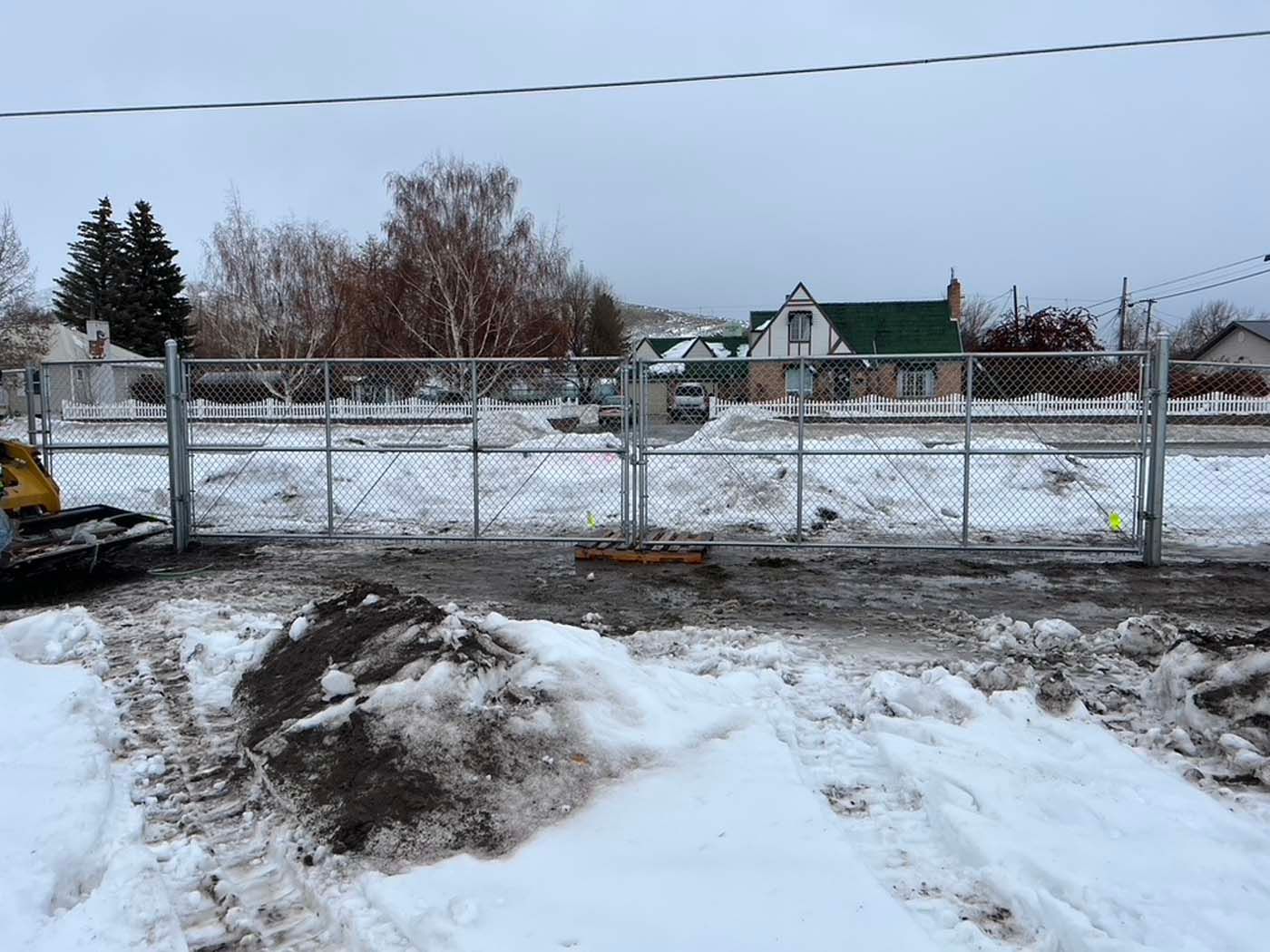 Snowy construction site with chain-link fence, muddy ground, and houses in the background