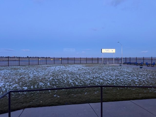 Snow-dusted grassy field behind a black fence at dusk, with a lit sign in the distance.