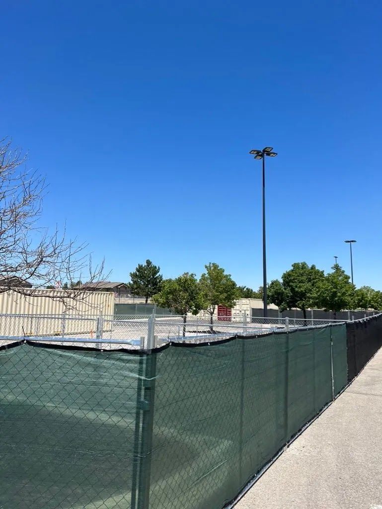 Construction fencing around a paved lot under a clear blue sky, with tall light poles and trees nearby