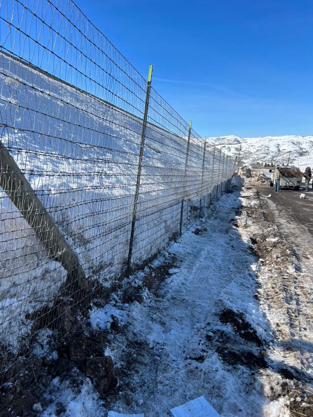 Snow-covered road beside a wire fence under a clear blue sky, with mountains in the distance.