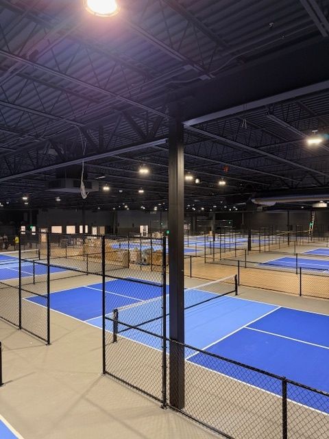 Indoor pickleball courts with blue playing surfaces and black netting under bright warehouse lights