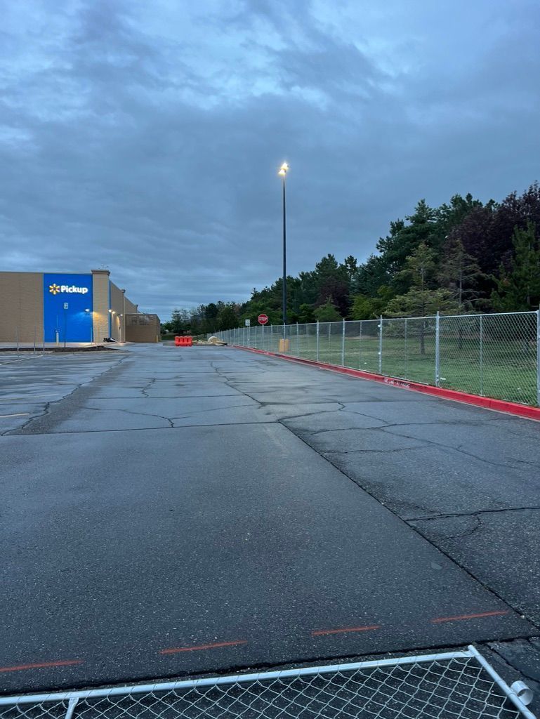 Empty parking lot at dusk with cracked pavement, a blue building, fence, and tall light pole under cloudy sky