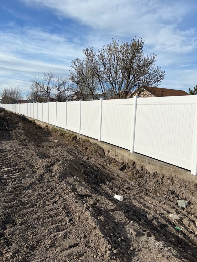 White vinyl fence along a dirt embankment beside bare trees under a cloudy blue sky.