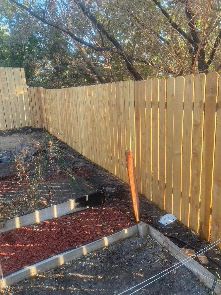 Wooden fence bordering a backyard garden bed with red mulch and a shovel leaning against it