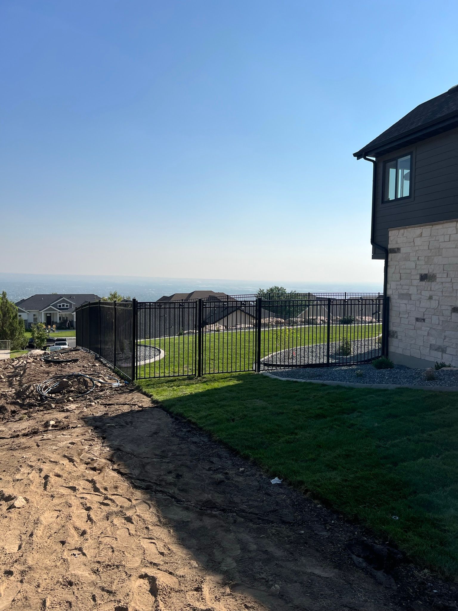 Backyard with dirt trench, black metal fence, green grass, and a stone house under a clear blue sky