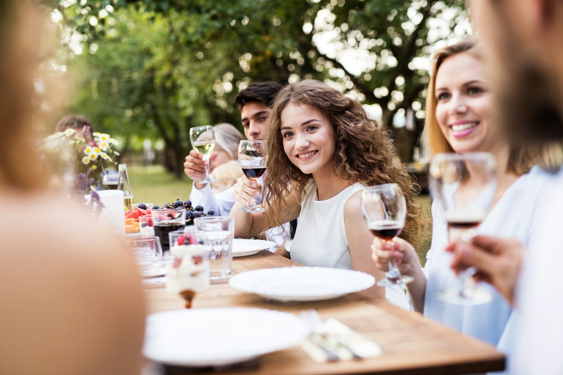 Friends dining outdoors at a table, smiling and toasting with drinks in a sunny garden.