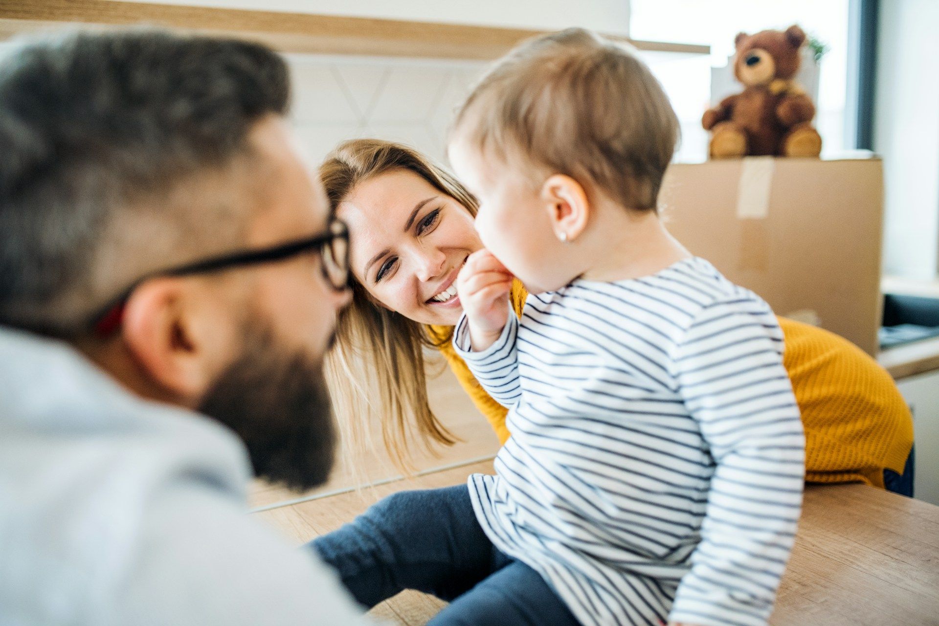 Adults smiling at a toddler indoors, with a teddy bear on a box in the background