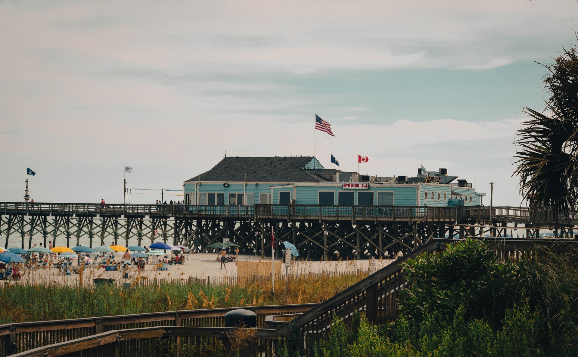 Beachfront pier with a building and American flag, viewed through dunes and greenery at sunset