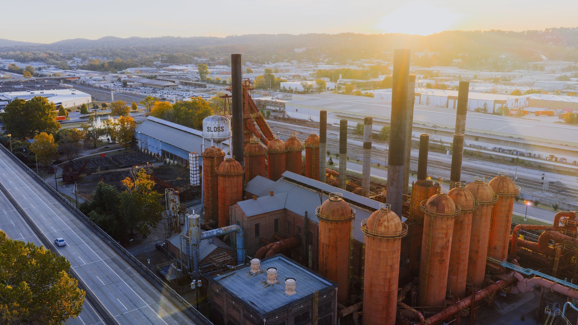 Sunlit rust-colored industrial plant with tall smokestacks beside a highway, overlooking a city