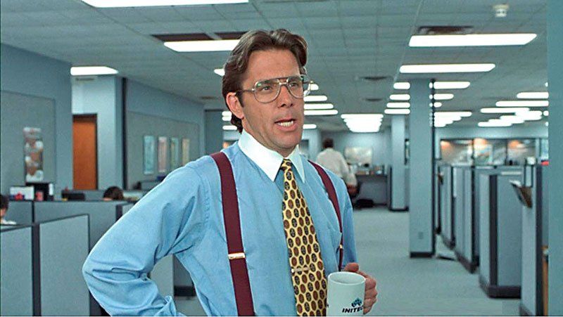 A man wearing suspenders and glasses is standing in an office holding a cup of coffee.