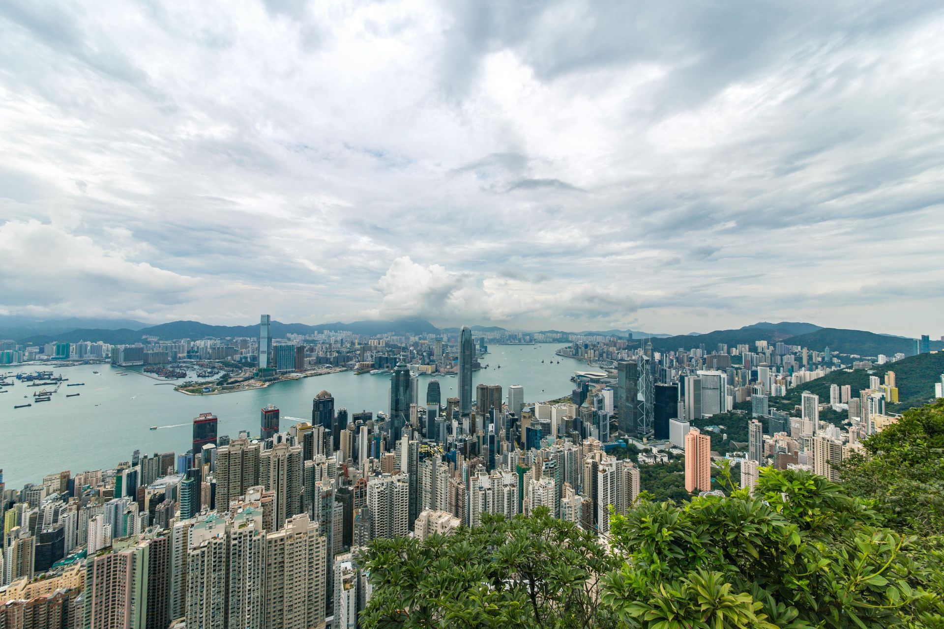 Hong Kong cityscape with skyscrapers, harbor, and cloudy sky viewed from a green, hilly area.