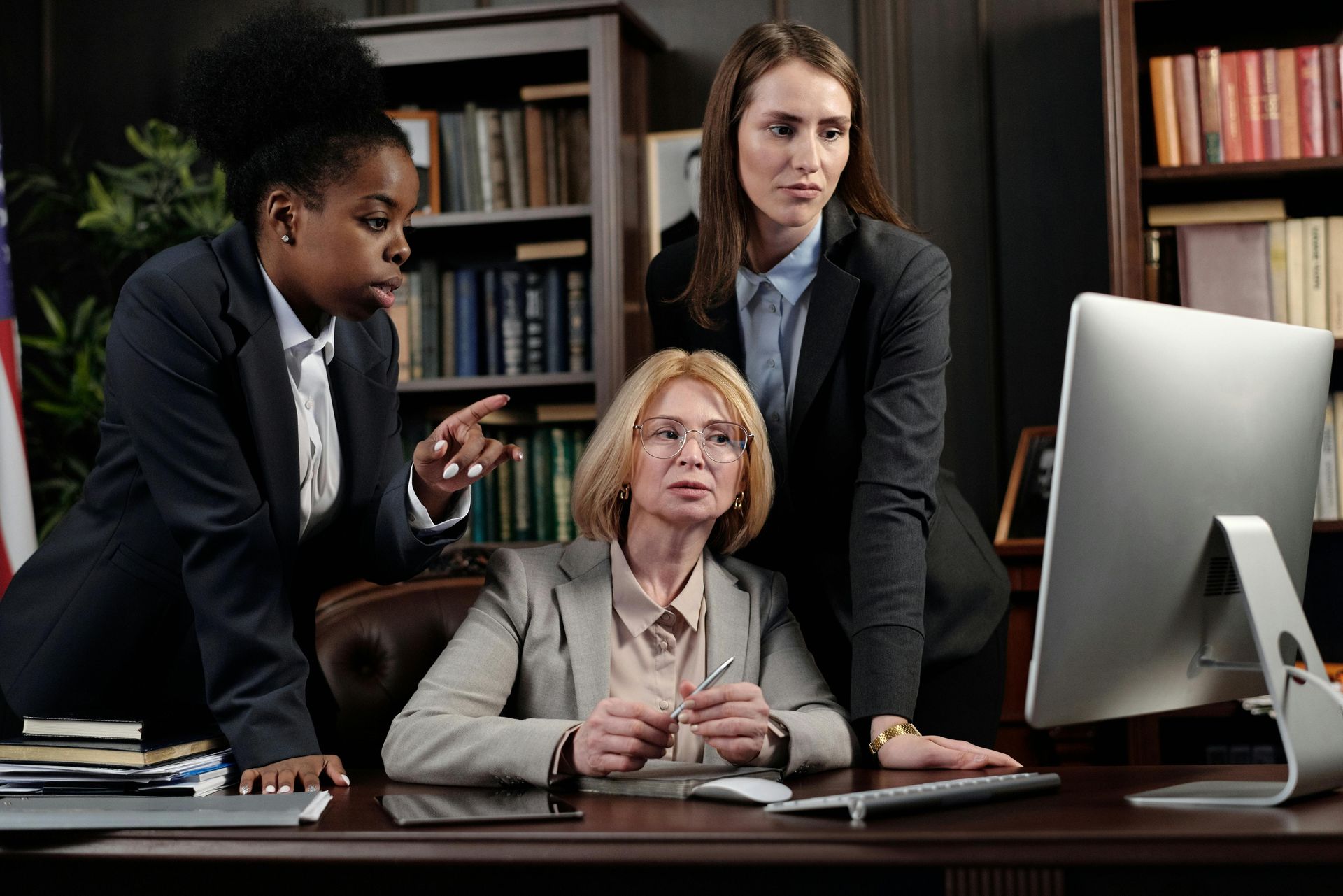 Three women in suits looking at a computer screen in an office. One points, others appear to be reviewing documents.
